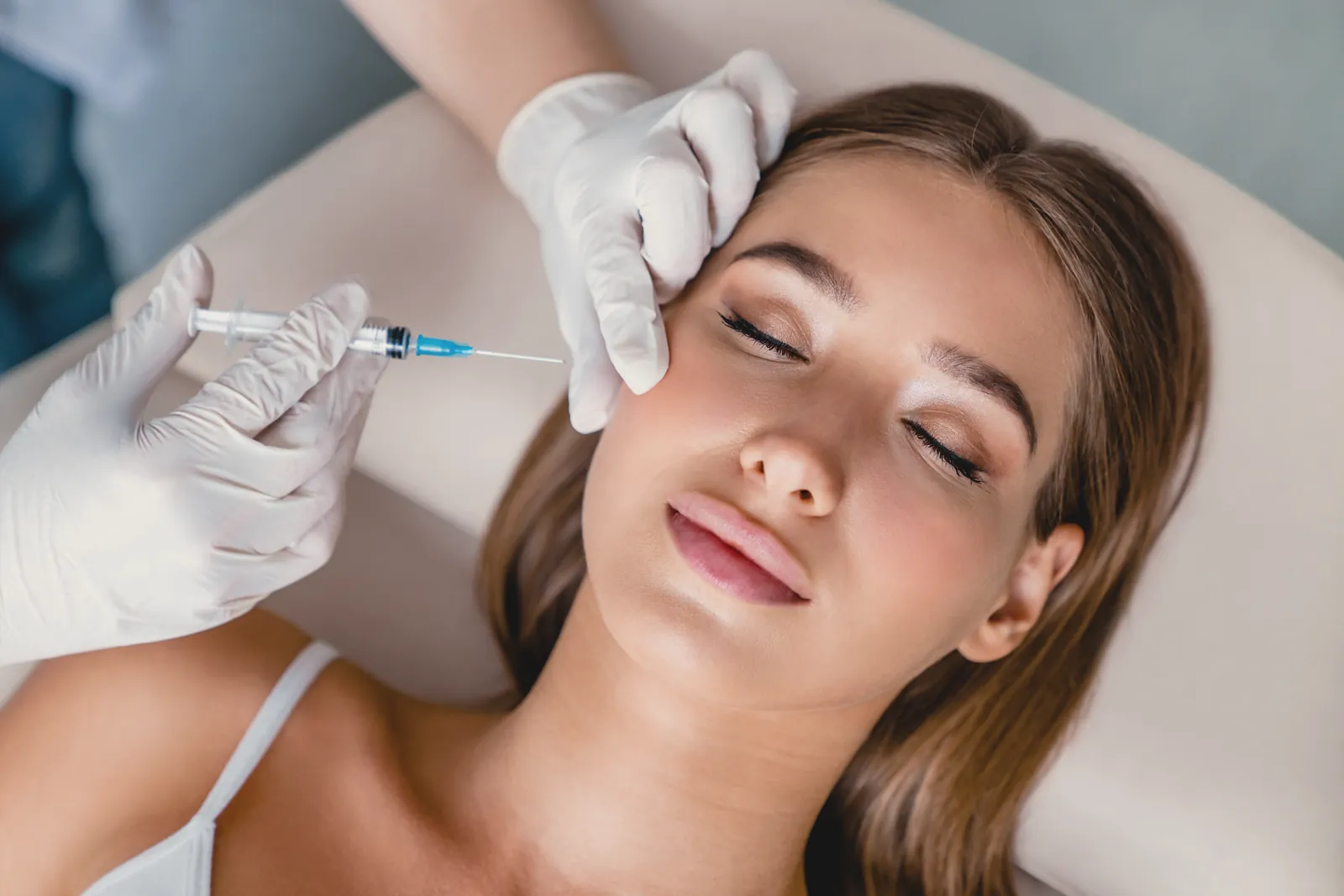 A gloved practitioner performs an injectable treatment near a relaxed client’s temple