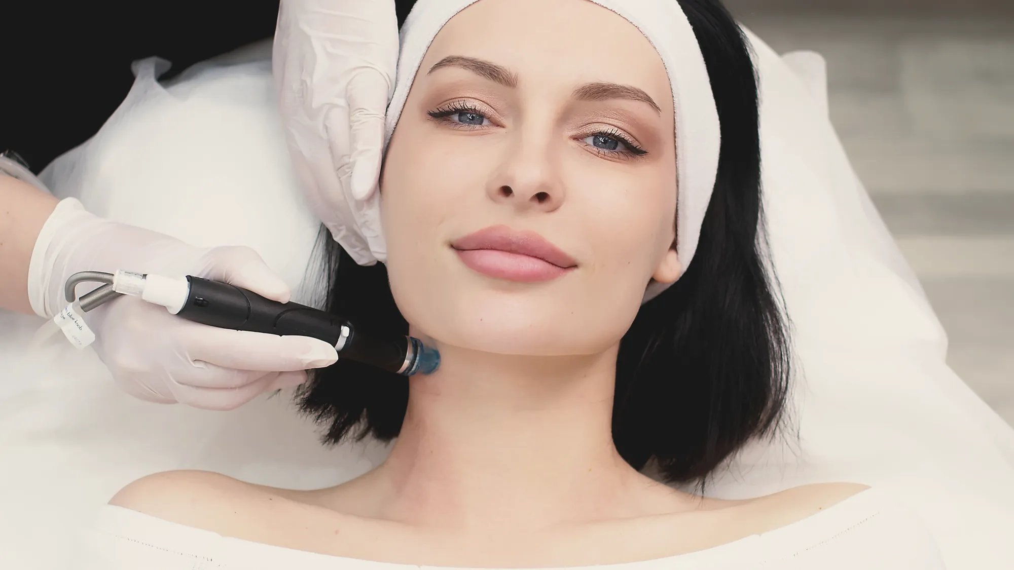 A gloved practitioner guides a handheld treatment device along a relaxed client’s jawline