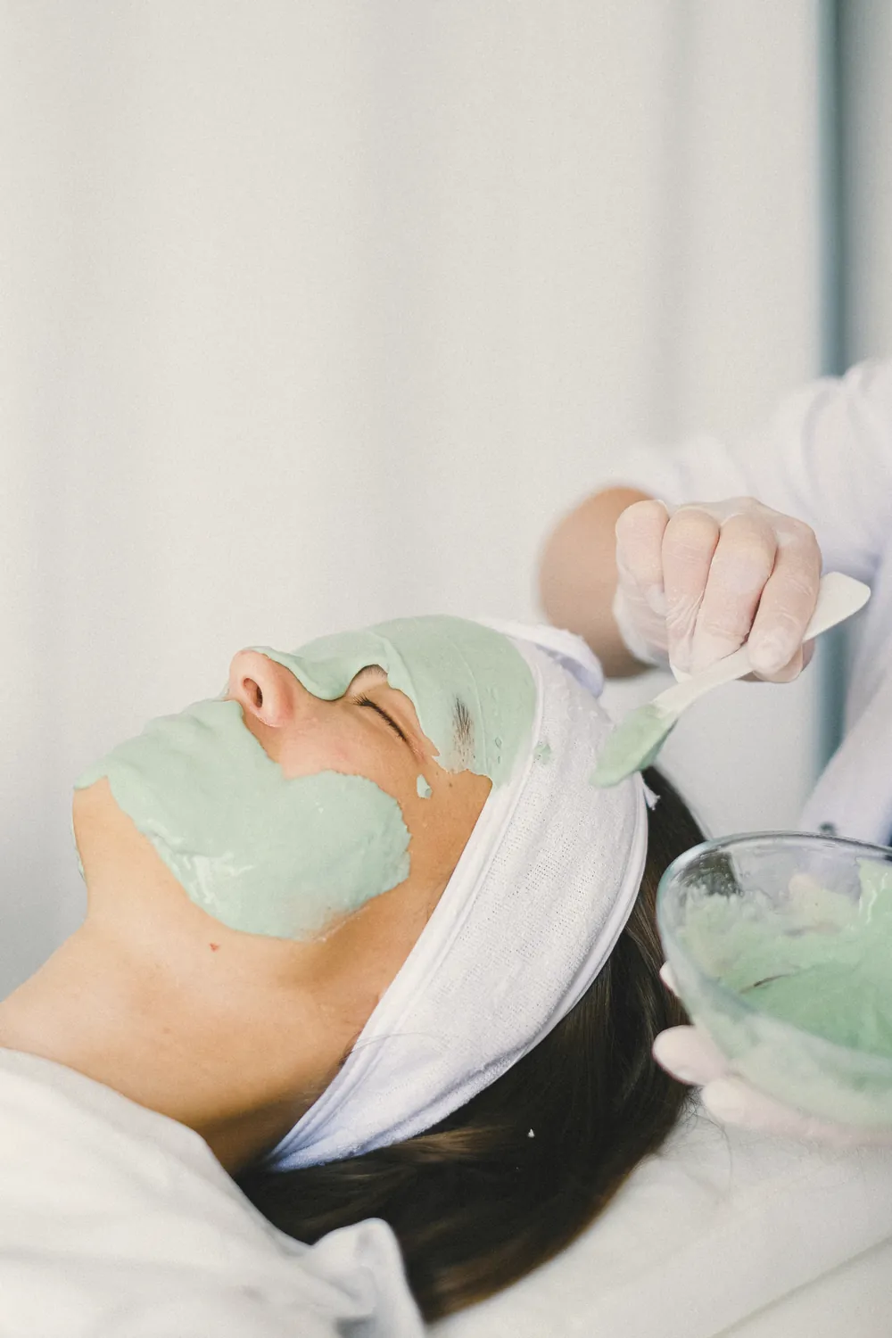 An aesthetician applies a soft green mask to a client’s face during a facial treatment