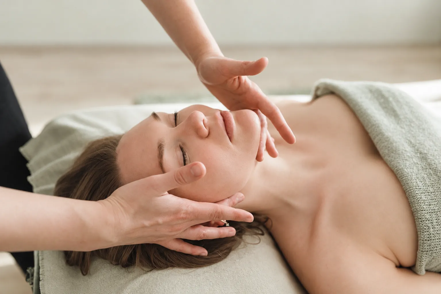 A relaxed client lies under a soft sage blanket as a practitioner gently cradles her face during a facial treatment